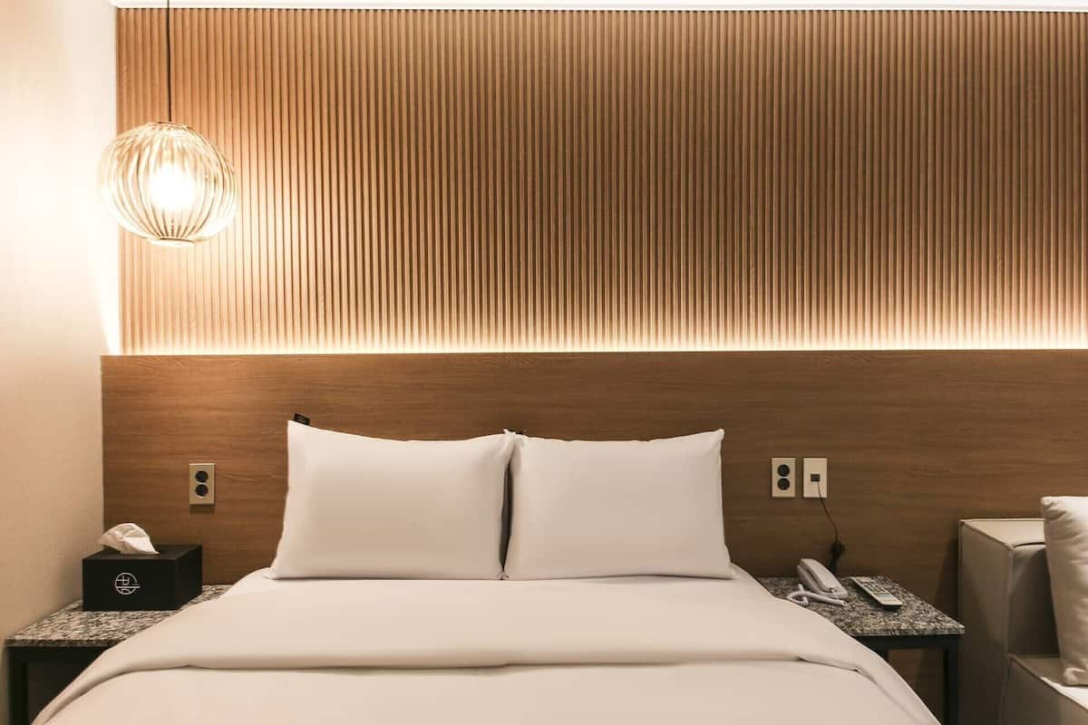A hotel room with a wooden headboard, a bed with white linens, a bedside table with a lamp, a telephone, and a remote control.