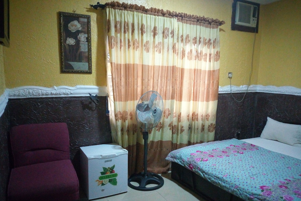 A small, compact room with a bed, a chair, a small refrigerator, and a standing fan.