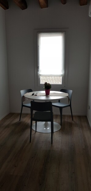 Een eetgedeelte met een ronde tafel, twee stoelen, een potplant en een raam met jaloezieën.