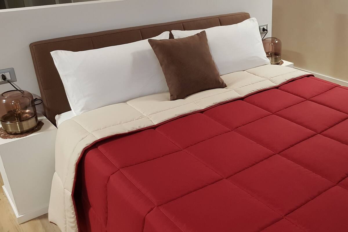 Un lit bien fait, avec une couverture rouge, des oreillers blancs et un coussin décoratif marron. Une serviette blanche pliée est posée sur le lit.