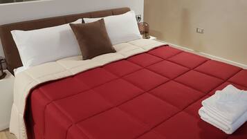 Una cama bien hecha con colcha roja, almohadas blancas y una almohada decorativa marrón. Hay una toalla blanca doblada sobre la cama.