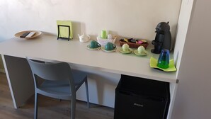 Een kleine kitchenette met een wit aanrecht, een zwarte prullenbak, een grijze stoel en diverse keukenbenodigdheden, waaronder een koffiezetapparaat, kopjes en een theepot.