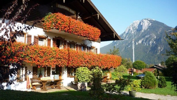 Ein traditionelles Chalet mit einem Balkon, der mit roten Blüten bepflanzt ist, einer Holzbank und einem Blick auf die Berge.