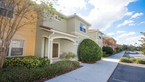 A residential building with a light-colored facade, a covered porch, and a well-maintained front yard with a hedge and shrubs.