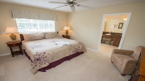 A bedroom with a bed, two bedside tables, a ceiling fan, a window with curtains, a chair, and a bathroom visible through an open door.