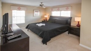 A bedroom with a bed, a desk with a computer and remote controls, a ceiling fan, and two bedside lamps.