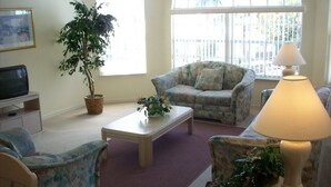 Ein Wohnzimmer mit Sofa, Sessel, Couchtisch und Lampe. Ein großes Fenster bietet Blick auf einen Zaun und einen Blumentopf.