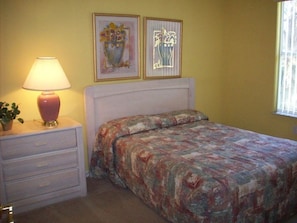 A bedroom with a bed, a bedside table with a lamp, two framed artworks on the wall, and a window with blinds. - Ip60570 - Villas at Island Club Lindfields - 3 Bed 2 Baths Condo (Kissimmee)