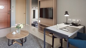 A modern hotel room with a wooden door, a round table with a vase of flowers, a desk with a lamp, a flat-screen TV, and a blue chair. - Jolley Hotel (Taipei)