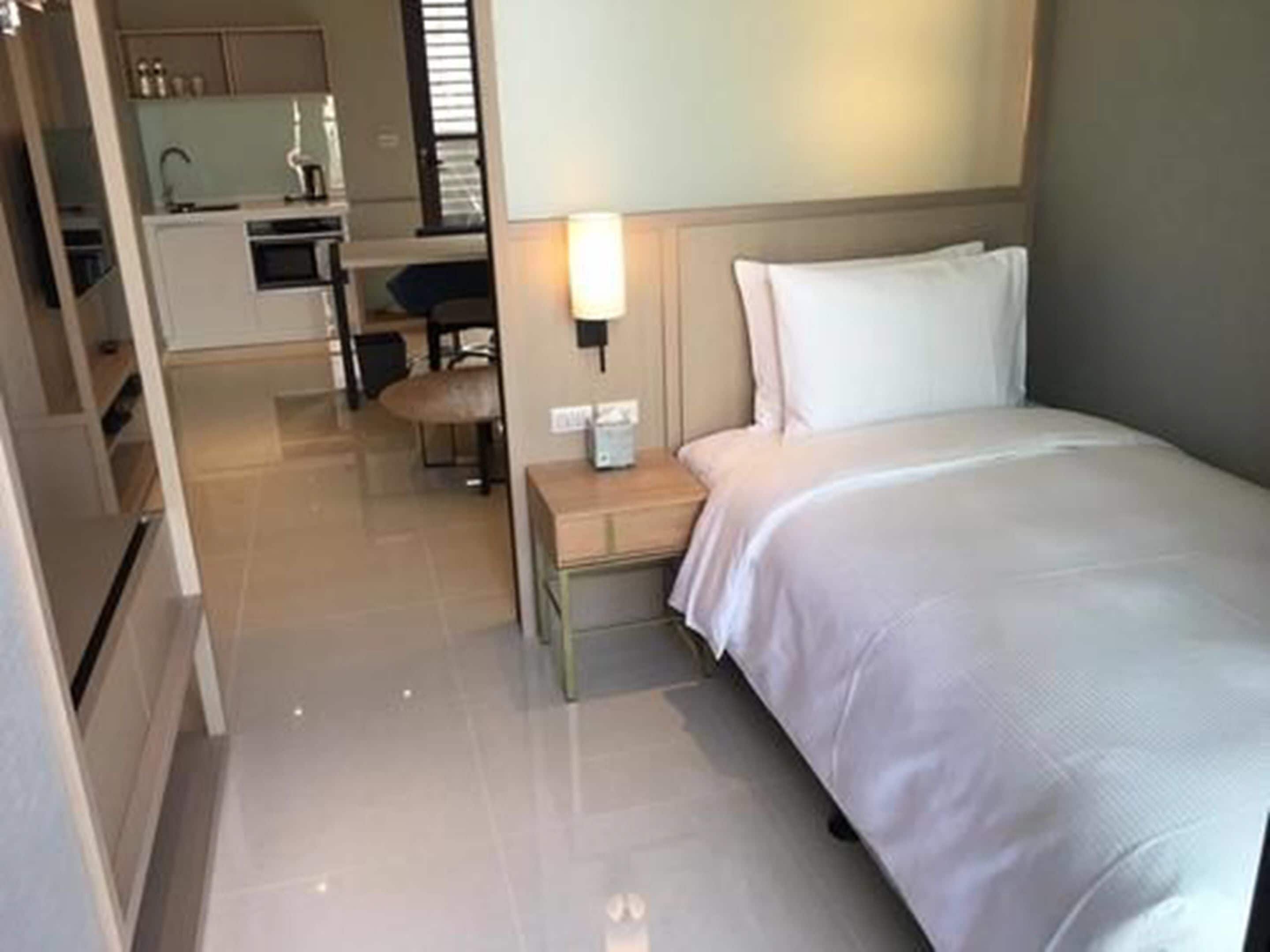 A hotel room with a bed, a bedside table, a lamp, and a view of the kitchen area.