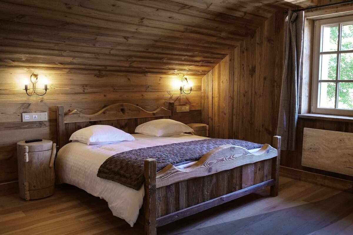 Une chambre dans une cabane en bois, avec un lit, deux lampes fixées au mur, une fenêtre avec des rideaux et un plancher en bois.