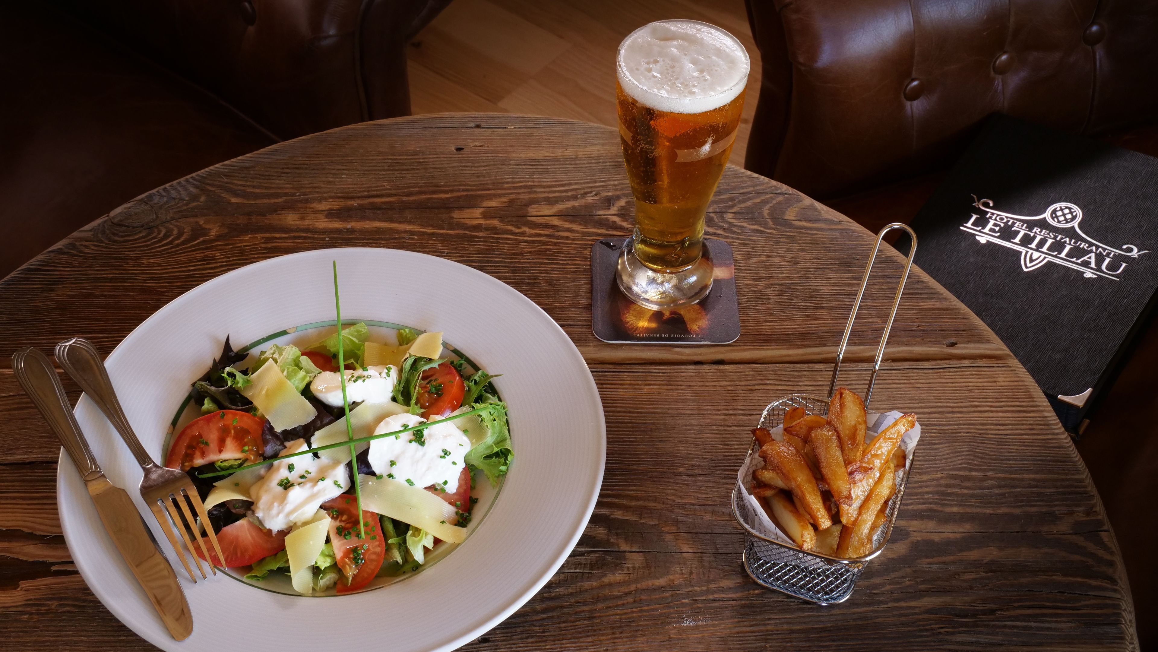 A wooden table with a plate of salad, a glass of beer, and a basket of fries.