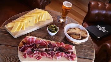 Une table en bois, sur laquelle on trouve une planche à fromages, un bol d’olives, un panier de pain et un verre de bière.