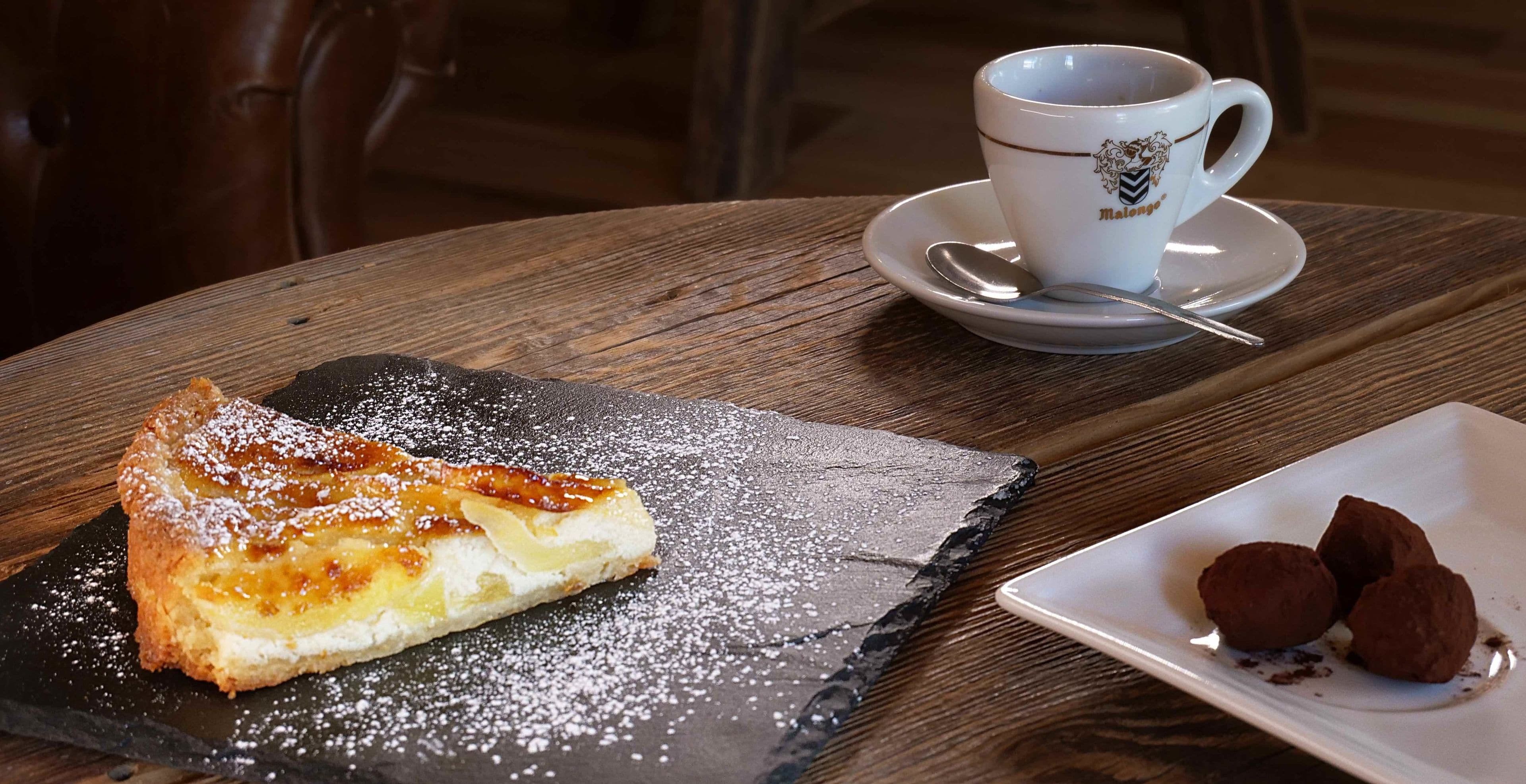 Une assiette à dessert avec une part de gâteau et du sucre glace, une tasse de café avec sa soucoupe, et une cuillère sur une table en bois.