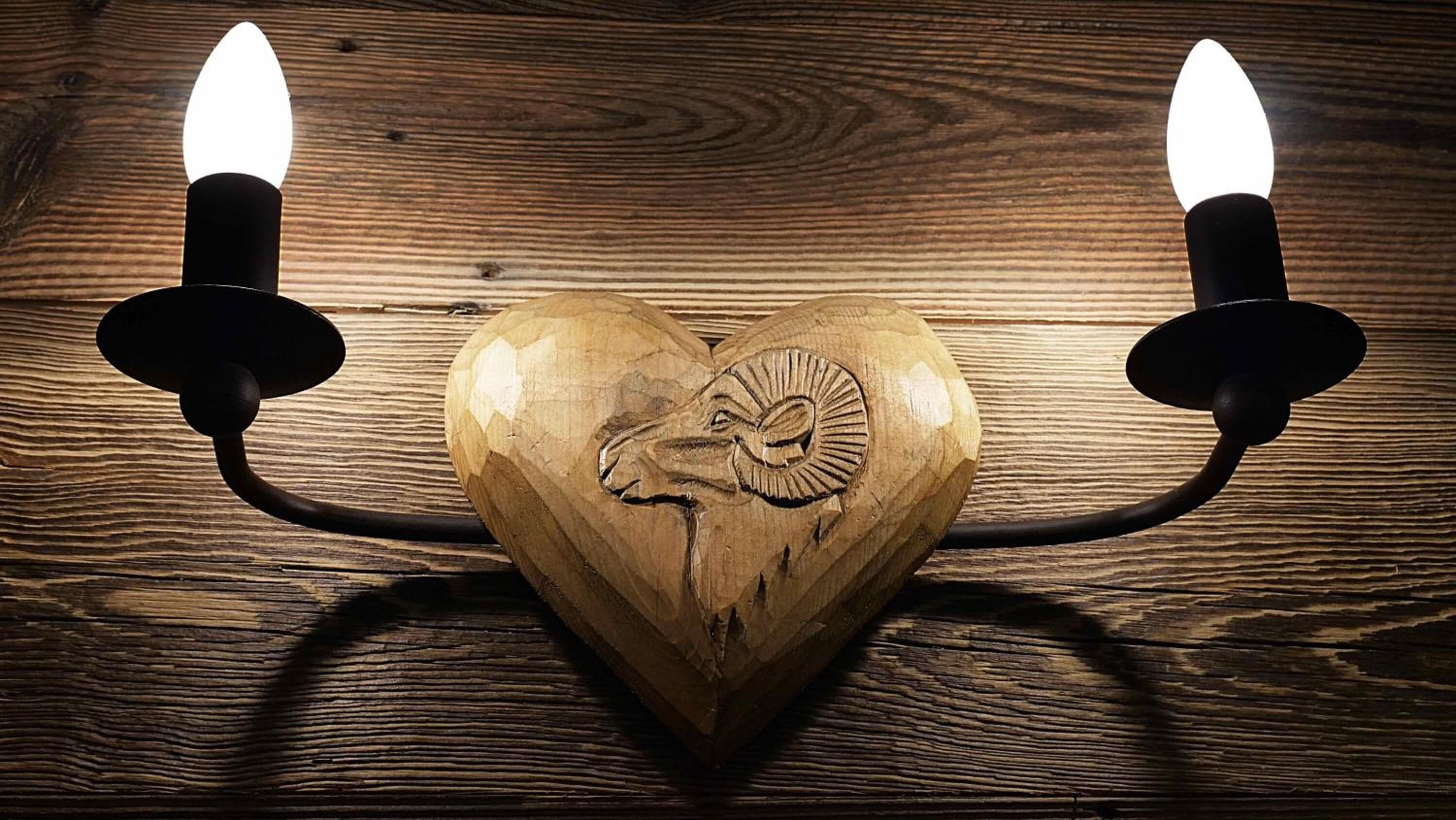Un candelabro de madera con forma de corazón, con cabeza de carnero tallada, dos velas y un fondo de madera.