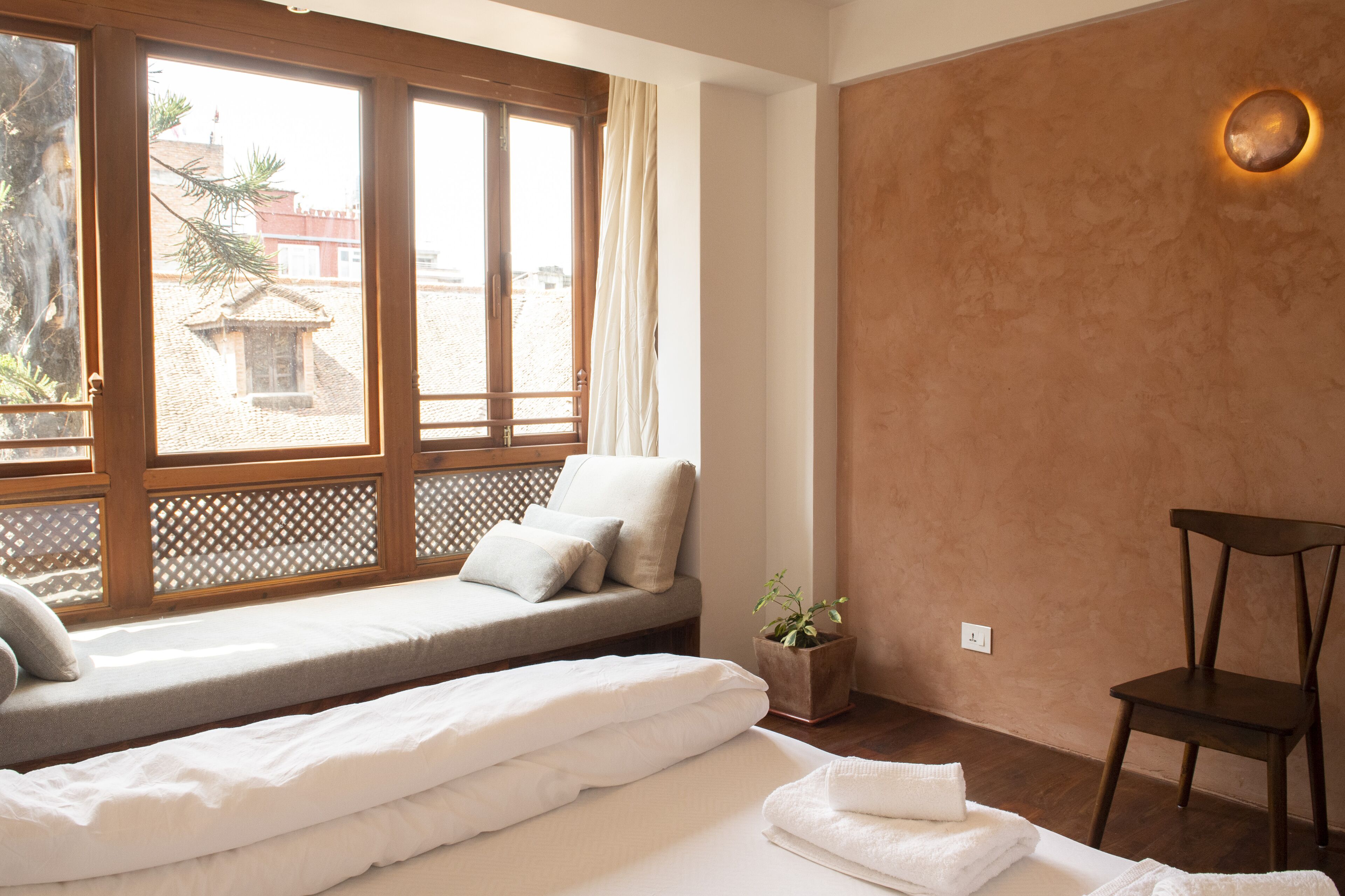 Quarto com uma cama, janela com vista para prédios, uma cadeira de madeira e um pequeno vaso de planta.