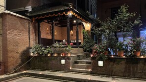 A traditional building with a tiled roof, brick walls, and a small porch area with steps leading up to it. There are potted plants and candles on the steps, and a tree is visible to the right. - Newa Chen Historic House UNESCO (Lalitpur)