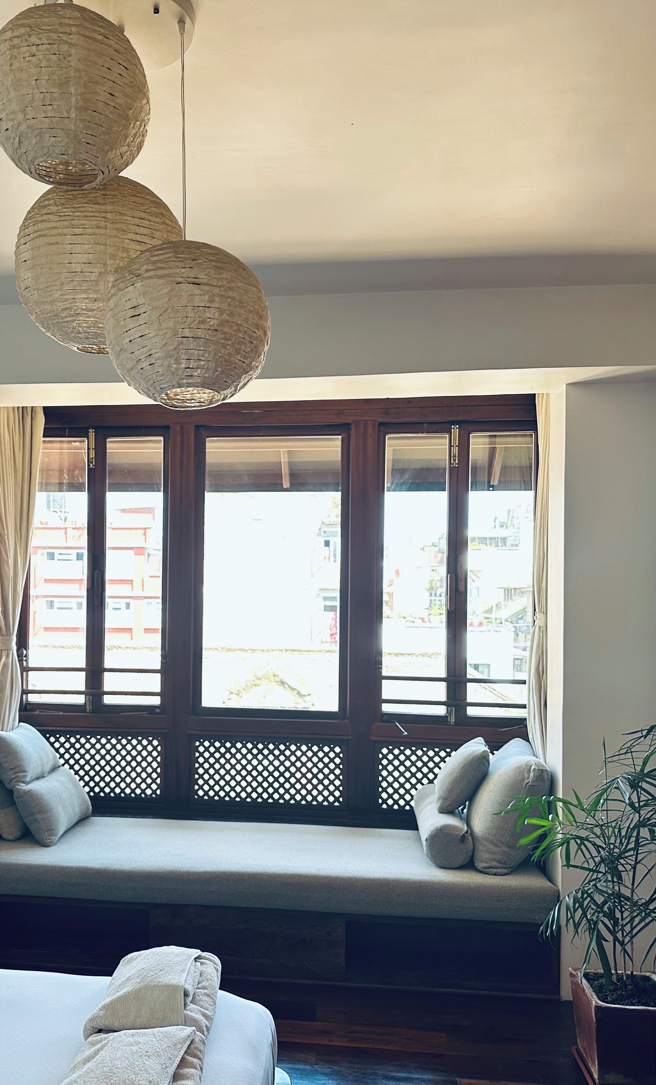 A room with a large window, a woven ceiling light fixture, a cushioned bench, and a potted plant.