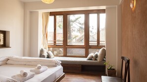A bedroom with a large window, a bed with white linens, a bench with cushions, and a wooden chair. - Newa Chen Historic House UNESCO (Lalitpur)