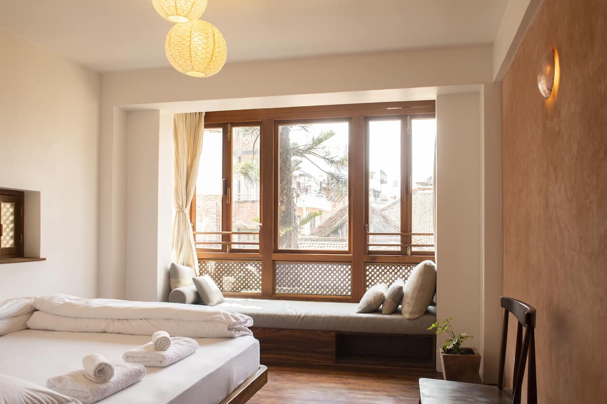 A bedroom with a large window, a bed with white linens, a bench with cushions, and a wooden chair.