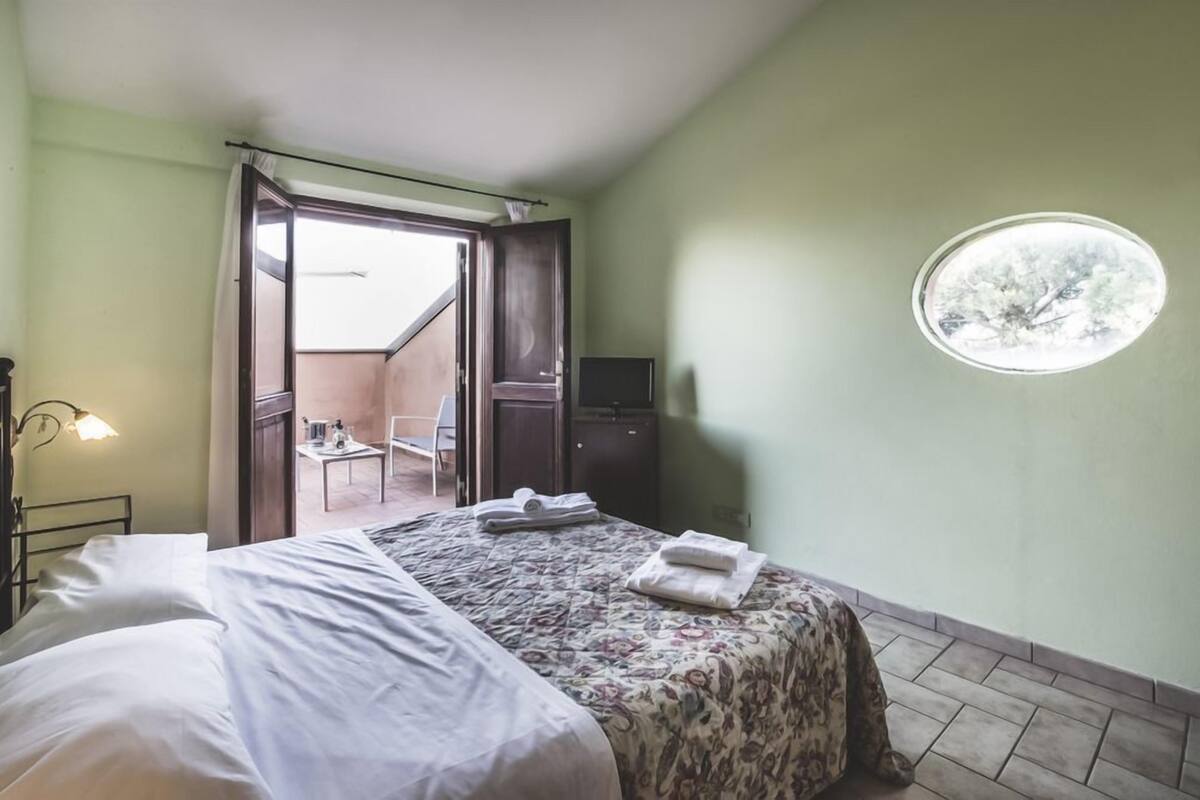 Un dormitorio con cama, una mesita con silla, televisión y ventana con vistas.