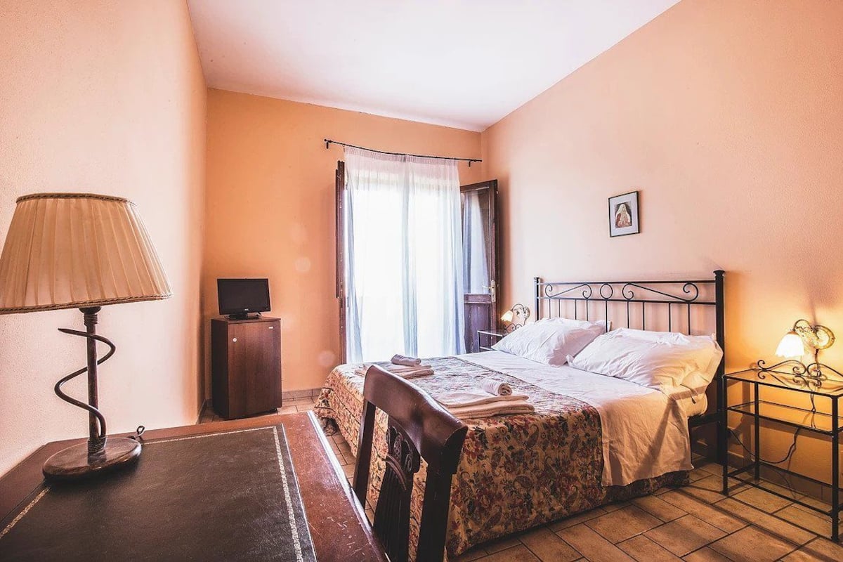 Un dormitorio con cama, escritorio, lámpara, televisión y ventana con cortinas.