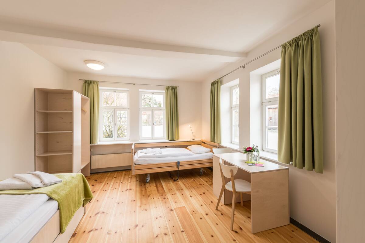 A compact room with a bed, a desk, and a chair. There are two windows with curtains, a radiator, and a shelf unit.