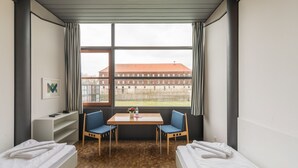 A room with two beds, a table, and chairs. There is a window with a view of a building outside. - Kloster Volkenroda (Körner)