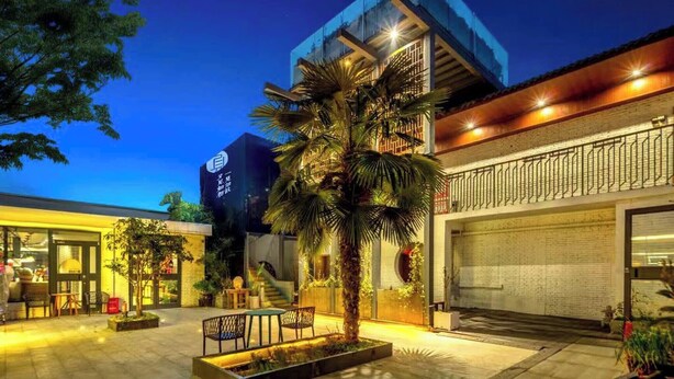 A modern building with a garage door, a small outdoor seating area with tables and chairs, and a palm tree. - First Sight Hotel,Next to Yingtianmen Imperial City,Provide Airport and High-Speed Rail Station Pickup (Luoyang)