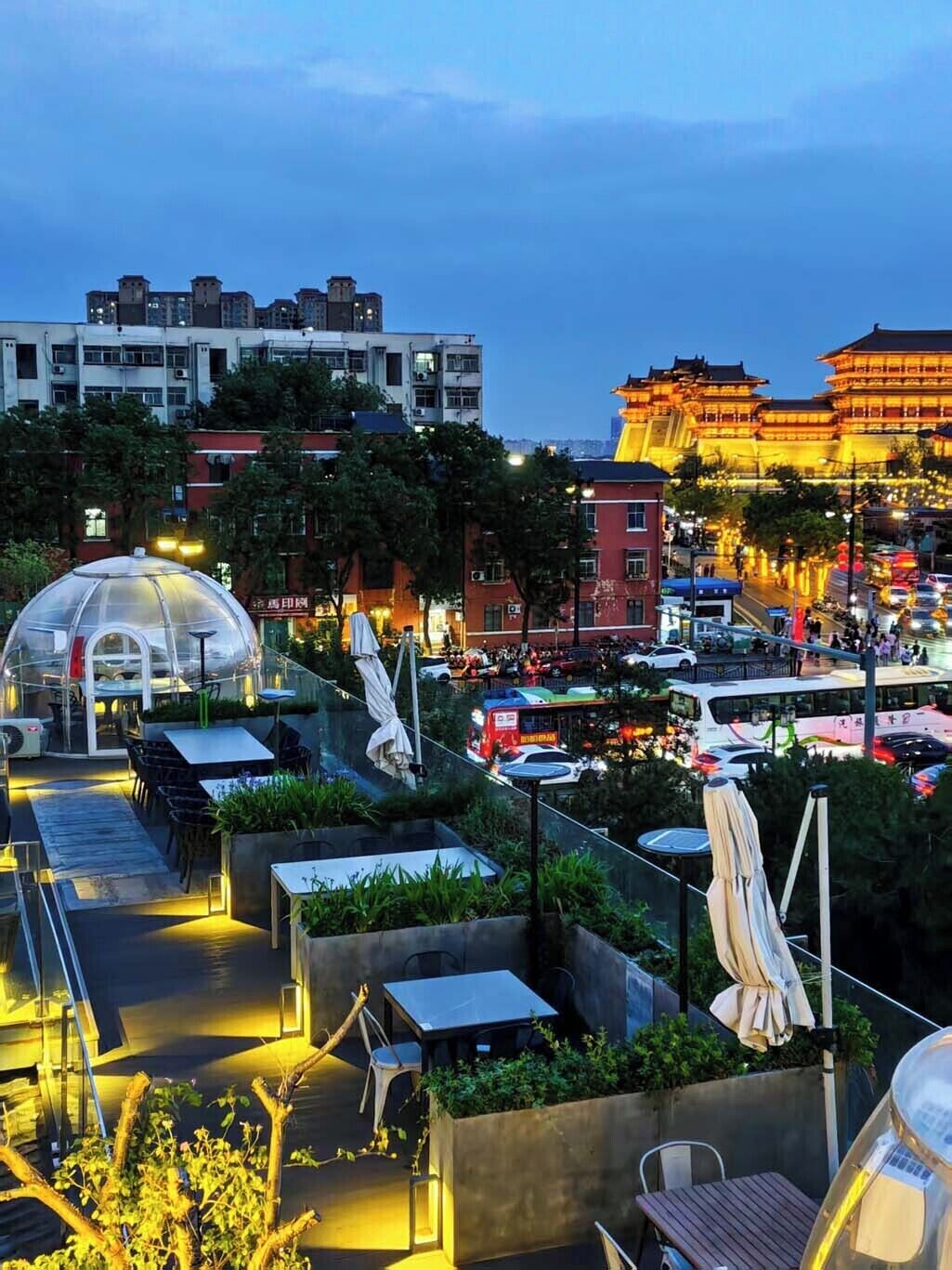 A rooftop terrace with outdoor seating, a glass greenhouse structure, and traditional architecture in the background.