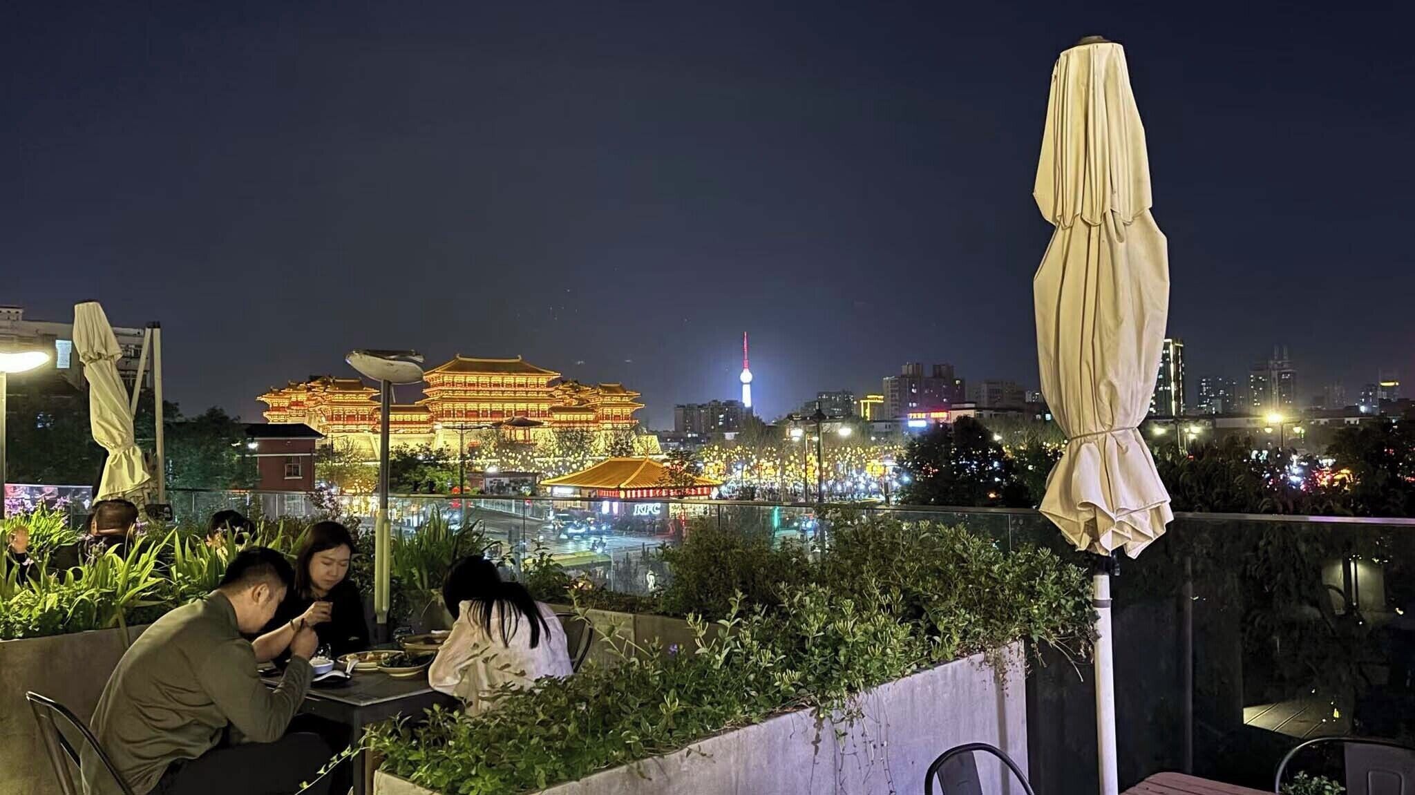 Un restaurant situé sur le toit d’un immeuble, avec des tables et des chaises, offrant une vue sur un bâtiment traditionnel dont l’architecture est mise en valeur par un éclairage nocturne.
