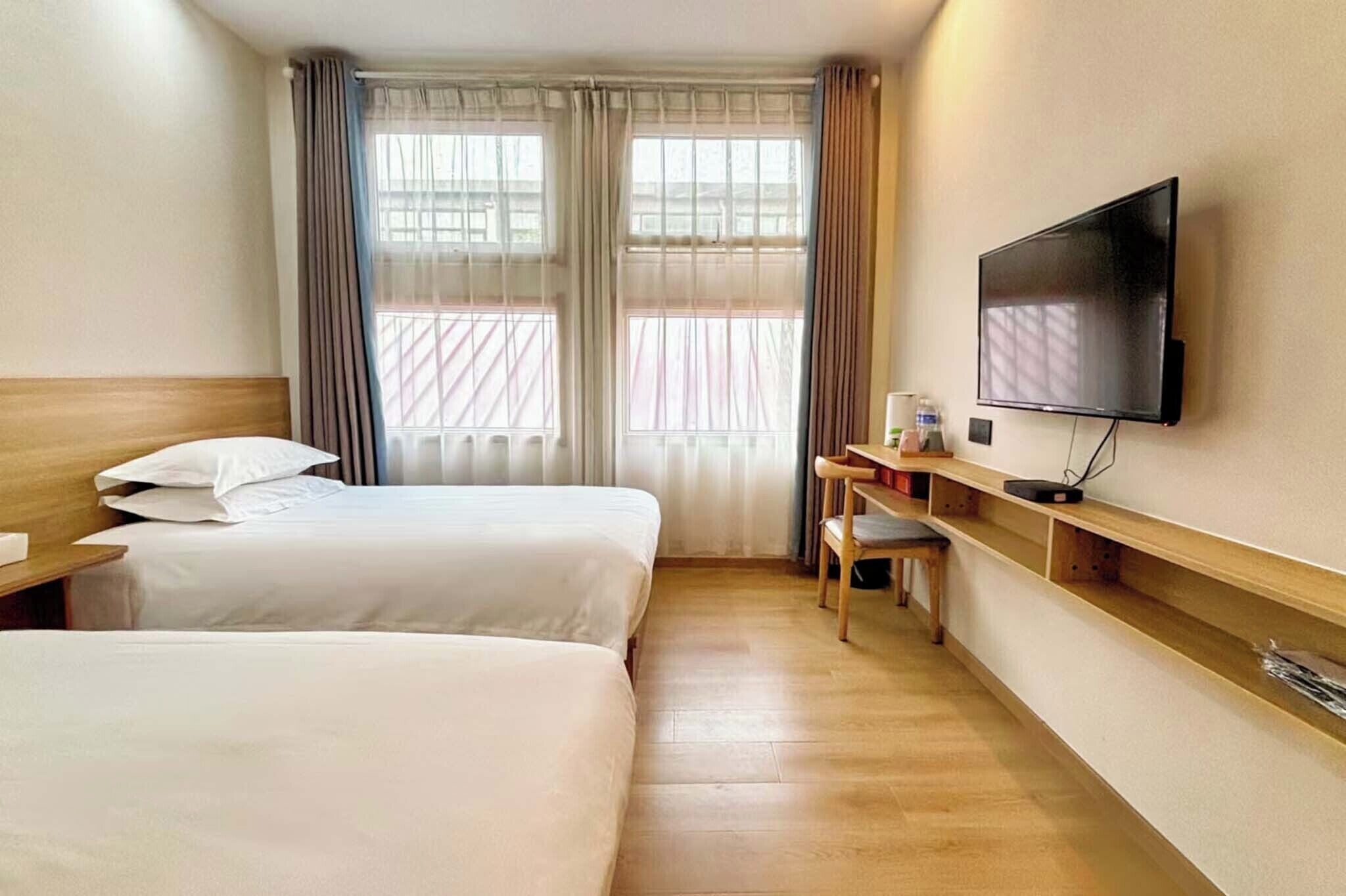 Habitación de hotel con dos camas, un escritorio con silla, televisor de pantalla plana montado en la pared y una ventana con cortinas.