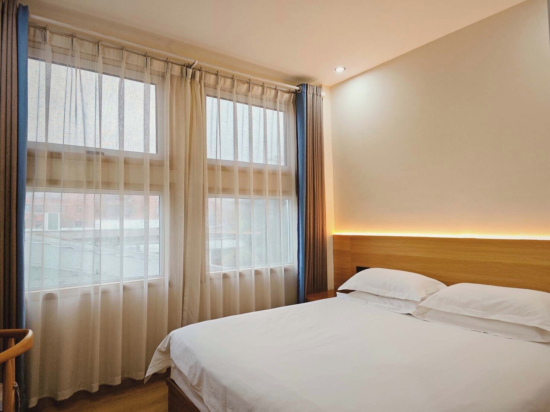 A hotel room with a large bed, two windows with sheer curtains, a wooden headboard, and a nightlight.