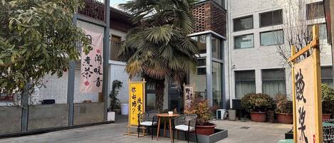 An outdoor seating area with tables and chairs, surrounded by potted plants and a palm tree.