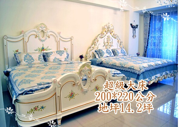 A bedroom with two beds, each with ornate headboards and floral bedding. The room has a large window with curtains.