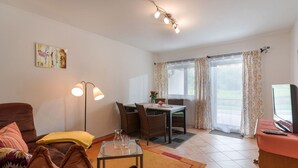 A living room with a brown sofa, a glass coffee table, a dining table with chairs, a TV on a stand, and a window with curtains. - Ferienwohnung Nr. 4 für 2 - 4 Personen 55 qm (Kiefersfelden)