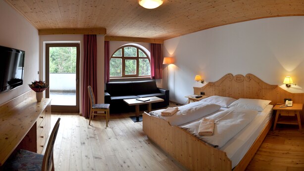 A wooden room with a bed, a sofa, a TV, and a window with curtains. - Berghotel Pine (Tires)