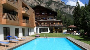 A pool area with lounge chairs and a wooden deck, a building with balconies, and a mountainous backdrop. - Berghotel Pine (Tires)