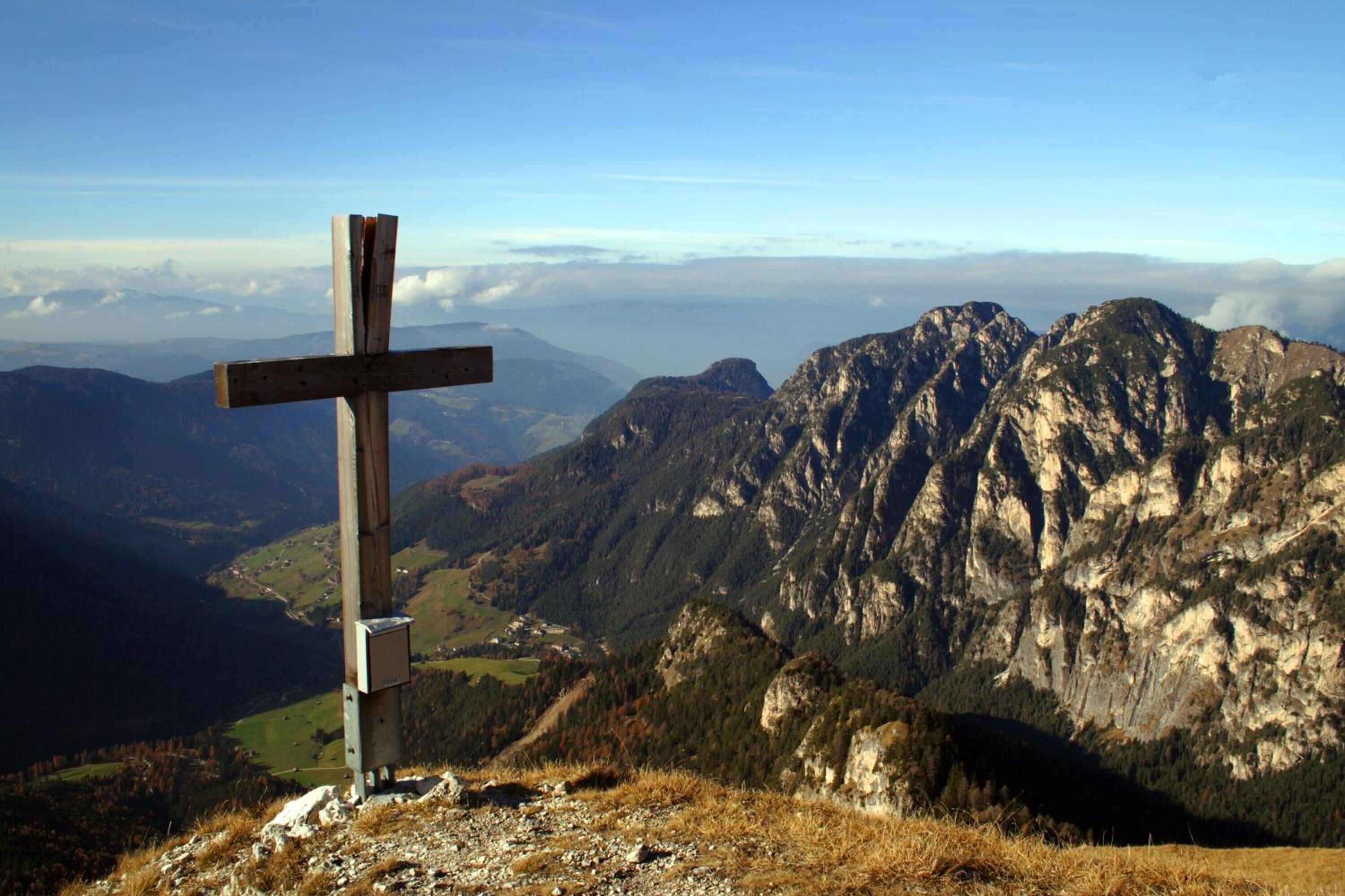 A wooden cross on a mountain peak with a view of rugged mountains and a valley below.