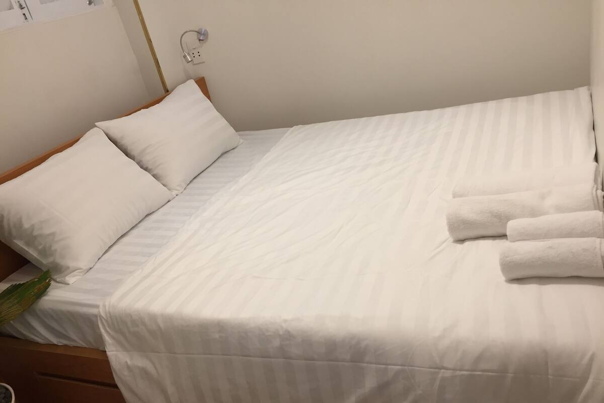 Un lit bien fait, avec du linge de lit blanc, une tête de lit en bois et une table de chevet sur laquelle se trouve une lampe.