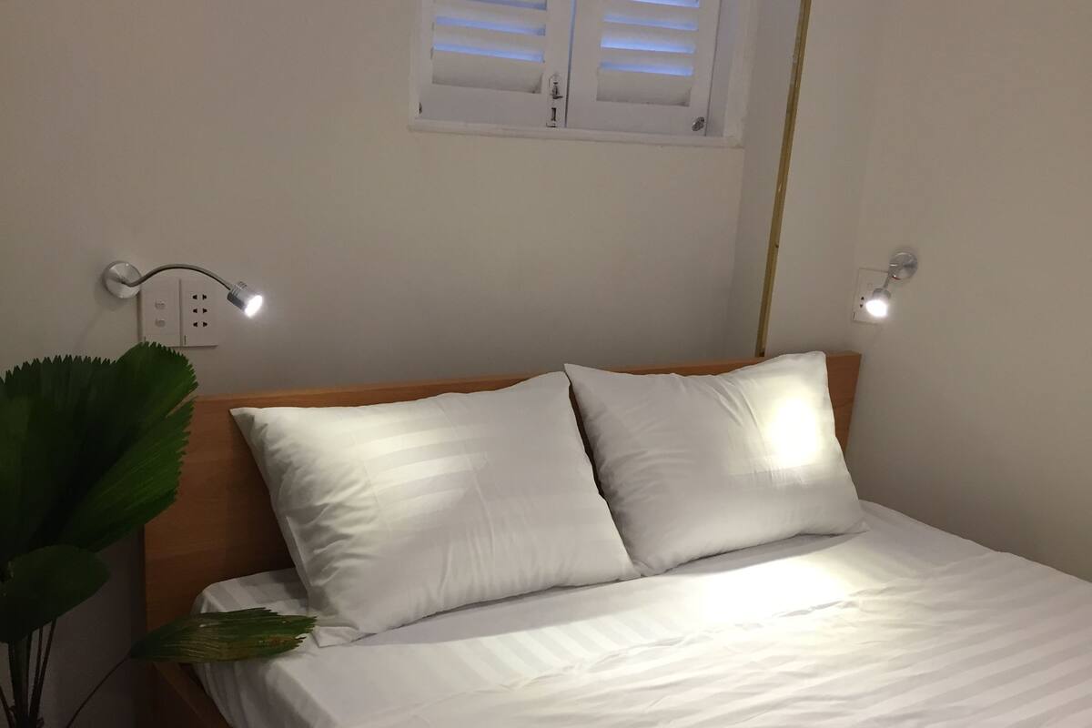 Un lit bien fait, avec du linge de lit blanc, se trouve dans une pièce dont la tête de lit est en bois. Une plante en pot est placée à gauche du lit, et on peut voir deux lampes fixées au mur.