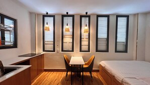 A compact room with a bed, a small table with two chairs, a kitchenette, and three windows with hanging lamps. - An Nam Corner Dreamer (Ho Chi Minh City)