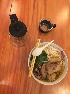 Un tazón de sopa de fideos con palillos y una cuchara, un frasco de vidrio y una taza pequeña sobre una mesa de madera.
