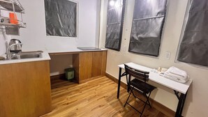 A compact kitchen with a sink, a small table, and a chair. There are two windows with blinds, and a green trash bin is visible. - An Nam Corner Dreamer (Ho Chi Minh City)