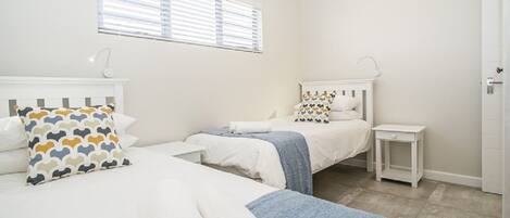 A bedroom with two beds, a window with blinds, a bedside table, and a door.