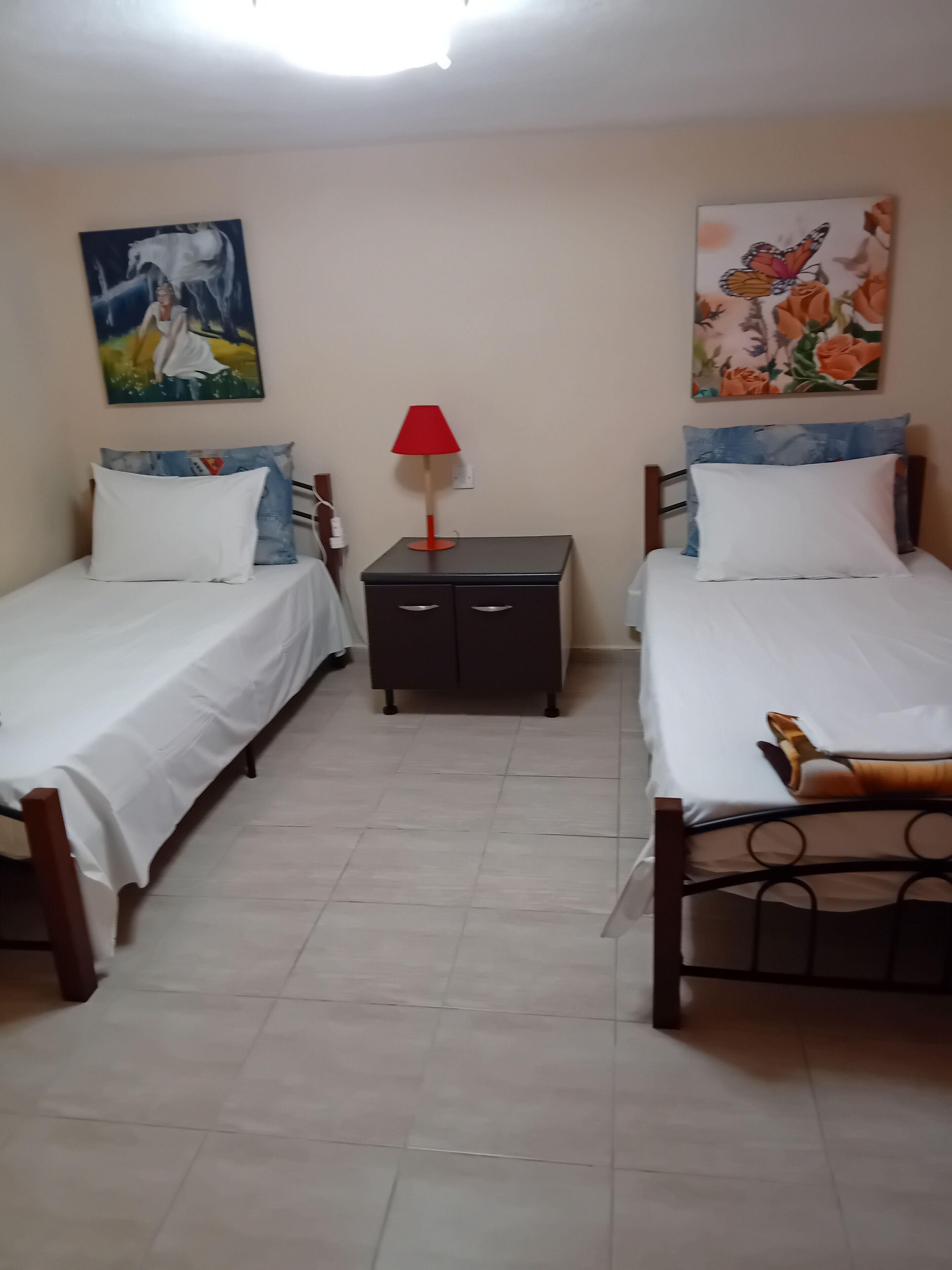 A room with two single beds, a nightstand, a red lamp, and two wall paintings.