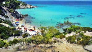 A beach with clear turquoise water, rocky cliffs, and sparse vegetation. - Maisonette Maria - for tranquility. Sykia Chalkidikis (Sithonia)