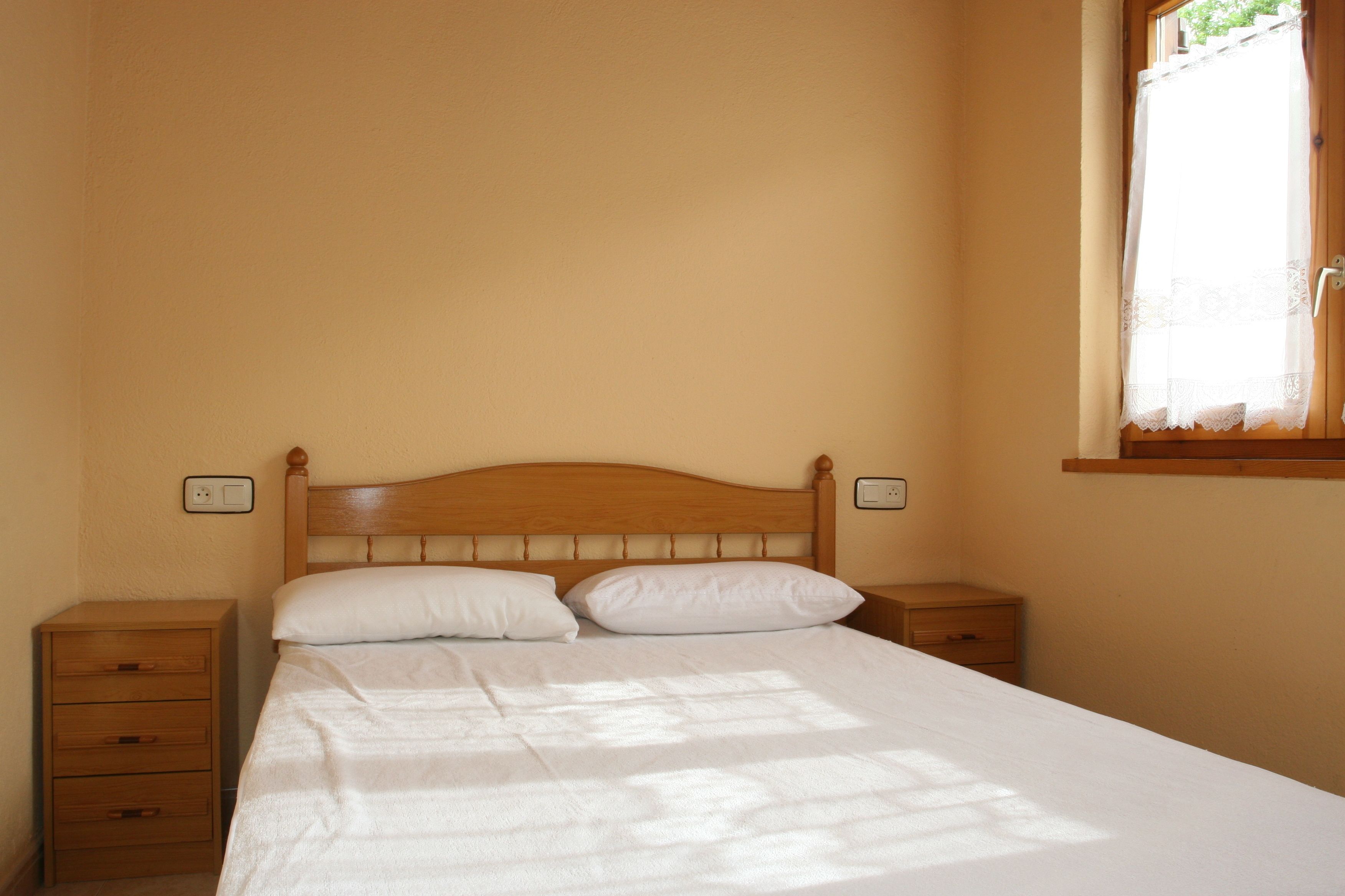 Ein Einzelbett mit Holz-Kopfteil, zwei Nachttische und ein Fenster mit weißem Vorhang.