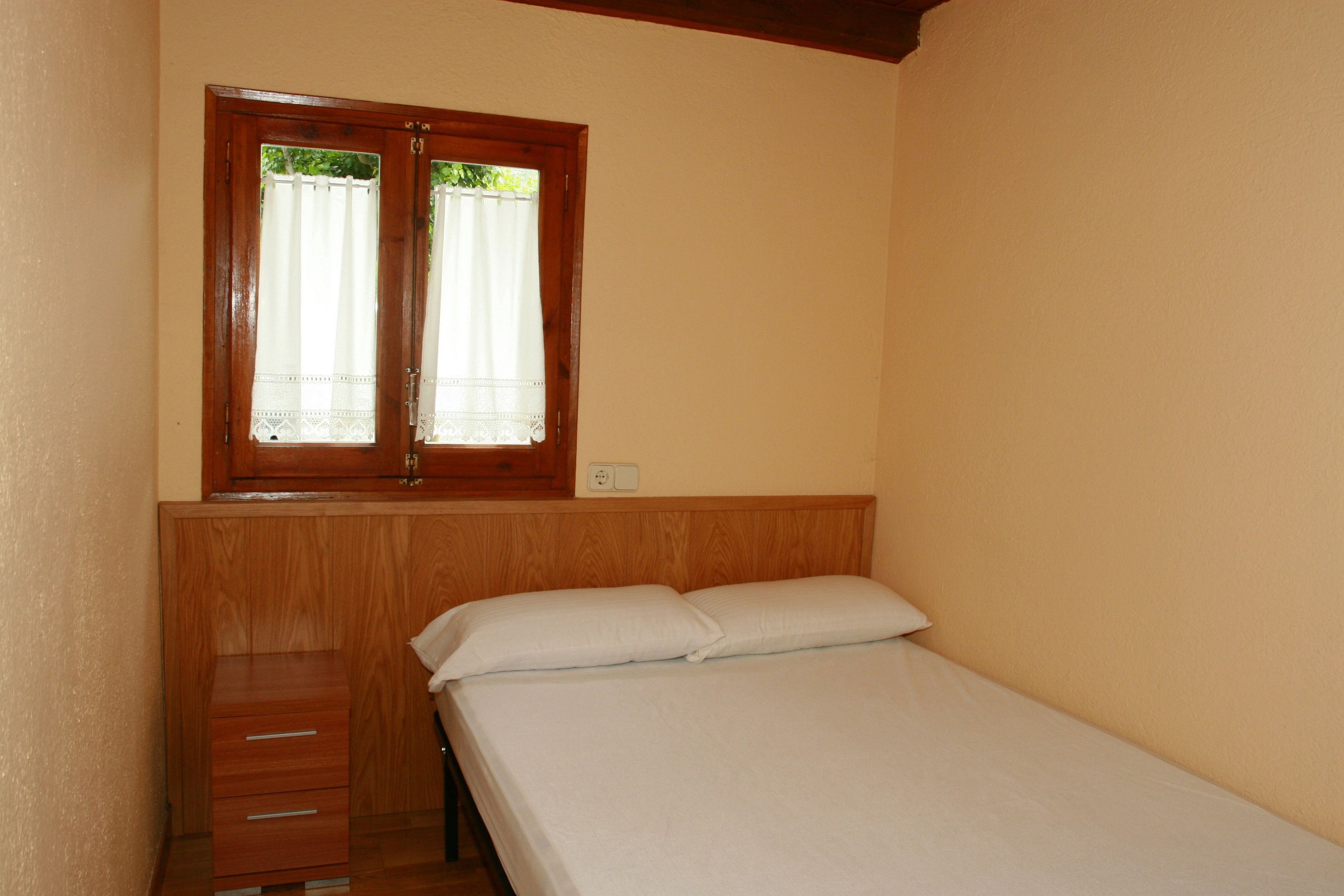 Una habitación pequeña con cama individual, cabecera de madera, ventana con cortinas blancas y un mueble de madera con cajones.