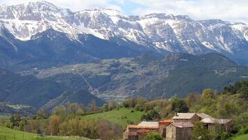 A rustic stone building with a sloped roof, surrounded by lush green fields and mountains in the background.